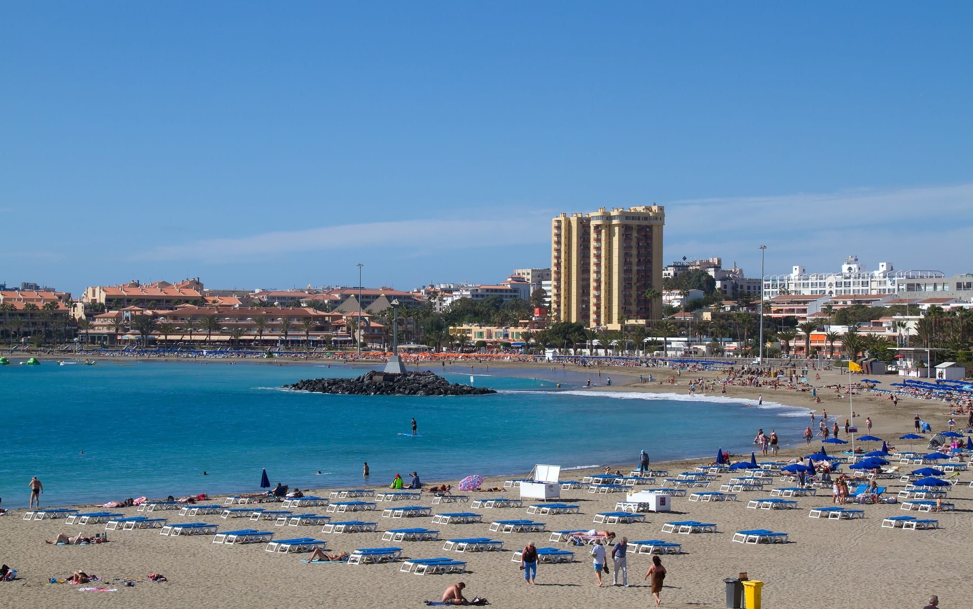 Playa de las Vistas in Los Cristianos, south Tenerife: rows of blue sunbeds on golden sand, a calm turquoise bay with a breakwater and seafront apartment blocks