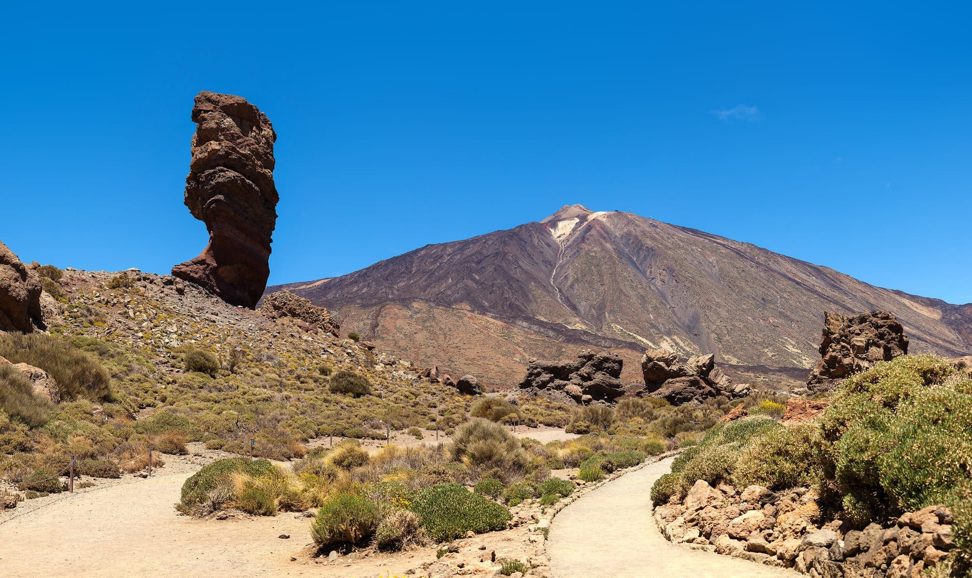 The Roque Cinchado rock pillar with Mount Teide behind, under a clear blue sky, a footpath winding through volcanic scrubland in Teide National Park